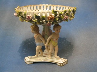 A circular 19th/20th Century Continental pierced porcelain table centre piece with basket work rim and floral encrusted decoration, supported by 3 cherubs, the base with crown shield mark 9"