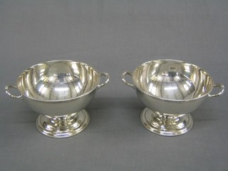 A pair of circular silver plated twin handled bowls with armorial decoration, raised on circular spreading feet