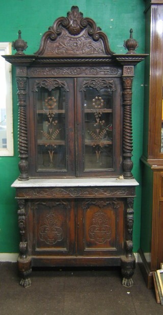 A 19th Century Continental carved wooden cabinet on cabinet, the upper section with arched carved back, the interior fitted shelves enclosed by glazed panelled doors above a white veined marble top, the base fitted a double cupboard enclosed by panelled doors, 37"