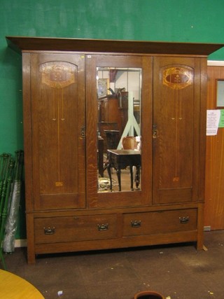 A handsome Arts & Crafts inlaid honey oak triple wardrobe with waisted cornice, the centre door fitted a rectangular bevelled plate mirror flanked by a pair of inlaid doors, the base fitted 2 long drawers 83" (silvering going to top left hand and bottom right hand of mirror, light contact marks by handles)