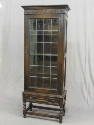 A 1930's carved oak display cabinet with moulded cornice, the interior fitted adjustable shelves enclosed by lead glazed panelled doors, the base fitted a drawer and raised on turned and block supports 24"