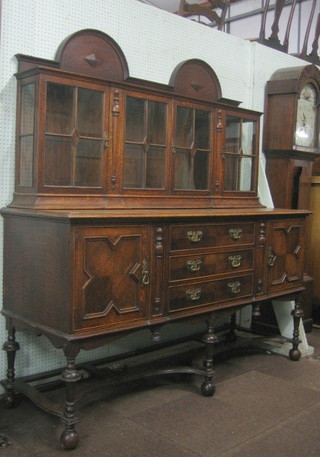 A 1930's oak Jacobean style sideboard with raised double domed back, fitted a display cabinet enclosed by  glazed panelled doors, the base fitted 3 long drawers flanked by a pair of cupboards, the base with X framed stretcher, raised on turned supports 70"
