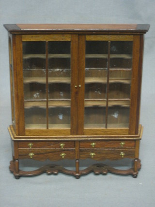 A dolls house mahogany finished display cabinet enclosed by glazed panelled doors, the base fitted 4 long drawers with wavy stretcher 7"