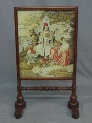 A Victorian mahogany fire screen with Berlin woolwork panel depicting a family scene, raised on bun feet
