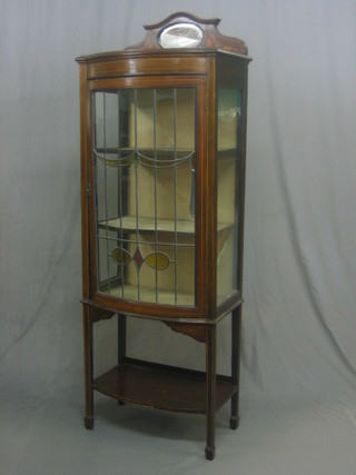 An Art Nouveau inlaid mahogany bow front display cabinet the raised back fitted an oval mirror, the interior fitted shelves enclosed by lead glazed panelled doors, raised on square tapering supports 24"