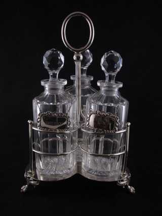 A silver plated 3 bottle decanter stand of triform shaped with 3 panel cut glass decanters, each with a silver plated decanter label   ILLUSTRATED