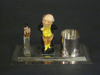 A rectangular silver plated 2 piece desk set comprising matching striker and cylindrical jar, flanked by a porcelain figure of Mr  Pickwick, by Hewkin & Heath