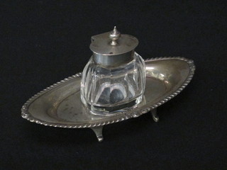 An Edwardian oval cut glass inkwell with hinged silver lid contained within a boat shaped silver tray, raised on panel  supports, Chester 1900