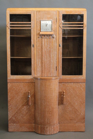 A 1930's Art Deco limed oak display cabinet on cabinet, the  upper section fitted a clock with square dial and Arabic numerals  flanked by a pair of cupboards enclosed by glazed panelled  doors, the base fitted a pair of cupboards enclosed by panelled  doors, raised on a platform base 36"