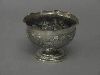 A circular Eastern embossed white metal bowl decorated figures, raised on a spreading foot