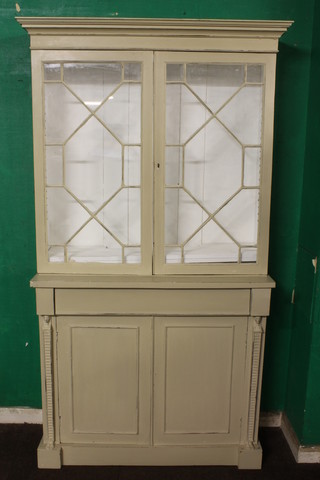 A late 19th Century cream painted bookcase, having a plain  moulded cornice above a pair of astragal bar glazed doors  enclosing 2 adjustable shelves above a single drawer with  cupboard doors below, flanked Egyptian style pilasters raised on  plinth base, 77"h x 45"w x 15"d