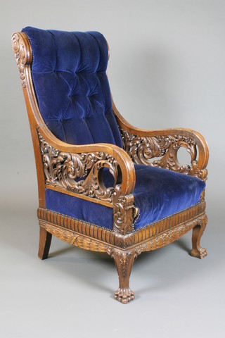 A late 19th Century carved oak library chair having foliate and scroll carved decoration, blue buttoned velvet upholstery, the base decorated with laurel and raised on braganza feet