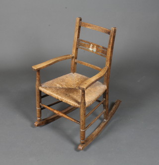 A late 19th Century Northern European stained beech wood childs rocking elbow chair, having ladder back, rush seat, on  turned legs and rockers