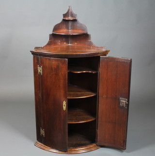 An early George III mahogany bow fronted hanging corner cupboard, cross banded, the upper section with 2 small shelves  above 2 cupboard doors enclosing 3 shelves with moulded base,  50"h x 27"w x 16"d