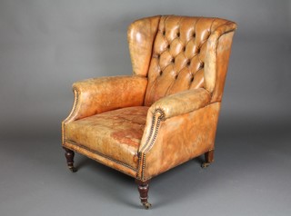 An Edwardian winged armchair upholstered in buttoned back brown leather, raised on turned supports with brass casters 