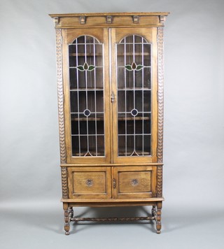 A 1930's carved oak display cabinet with moulded cornice and with carved panels to the sides, fitted shelves enclosed by lead glazed panelled doors, raised on spiral turned and block supports with H framed stretcher 39"h x 40"w x 15"d