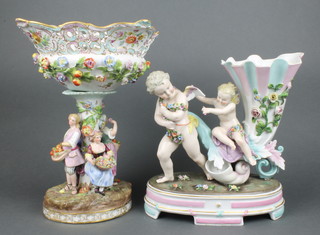 A late 19th Century German porcelain centrepiece in the form of a cherub pulling a cornucopia ridden by a fairy on a raised base 11", a ditto German centrepiece applied with flowers, the base with 4 figures 12"  