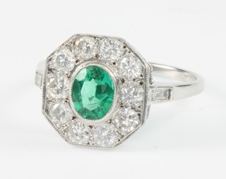 A white gold emerald and diamond cluster ring, the oval centre stone approx. 0.65ct surrounded by 10 brilliant cut diamonds with baguette shoulders, approx. 0.75ct, size N 1/2 