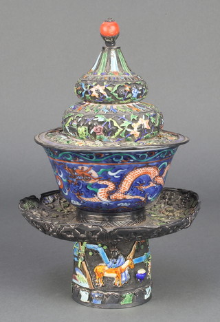 A late 19th/early 20th Century Chinese Tibetan silver enamel and repousse tea bowl holder, the signed base with a band of figures and animals in landscape, the lip decorated with flowers, the lid decorated with geometric and floral enamels, having a coral knop together with an early 20th Century Chinese blue ground tea bowl decorated with dragons, 8 1/2"
