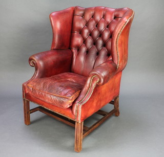 A Georgian style winged armchair upholstered in red button back leather raised on square supports with double H frame stretcher 