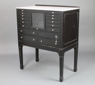A black painted dentists cabinet with white ceramic top, having a central cupboard with fitted interior flanked by 12 short drawers above 2 long drawers, raised on square tapering supports ending in spade feet 111cm h x 92cm w x 51cm d