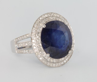 A 14ct white gold oval blue gem set dress ring approx. 6.5ct, surrounded by brilliant cut diamonds approx. 1.1ct in an open shank, size M 1/2