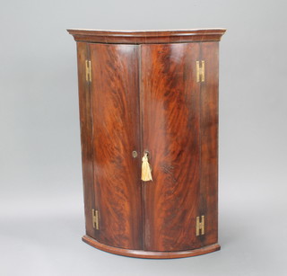 A Georgian mahogany bow front hanging corner cabinet with moulded cornice, the interior fitted shelves enclosed by panelled doors with brass H framed hinges 100cm h x 70cm w x 49cm d 