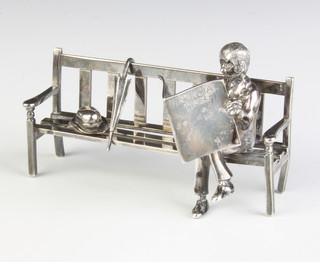 A silver model of  gentleman sitting on a bench reading  The Financial Times newspaper with his bowler hat and umbrella, London 1982, maker Sarah Jones, 70 grams 