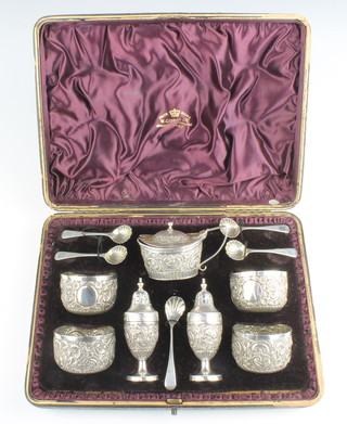 A Victorian repousse silver 7 piece condiment with scroll and floral decoration and 5 matching spoons, London 1889, maker George Maudsley Jackson, cased, 270 grams