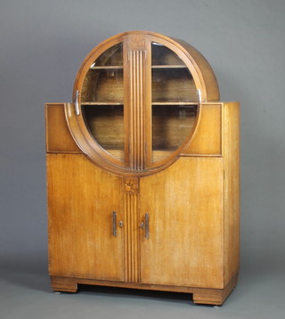 A 1930's Art Deco circular oak display cabinet on cabinet, the upper section fitted shelves enclosed by bevelled plate glazed doors the base fitted a cupboard enclosed by panelled doors 163cm h x 112cm w x 48cm d