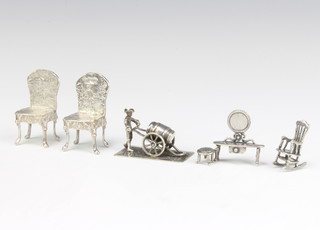 A pair of miniature Continental repousse silver high backed chairs 5cm, rocking chair, dressing table and a figure pushing a barrel, 90 grams
