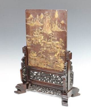 A second half of the 19th Century soapstone table screen, the chocolate-coloured stone carved with four houses built on rocks by a lake under pine trees and with details including a farmer with a buffalo, others in a rice field, mountains in the distance isolated in toffee-coloured inclusions, signed on the reverse 33.5cm x 26.5cm  contained in a carved hardwood stand  total height 50cm 