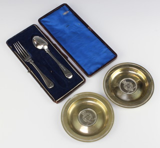 A pair of silver plated commemorative coin set dishes and a ditto christening spoon and fork 