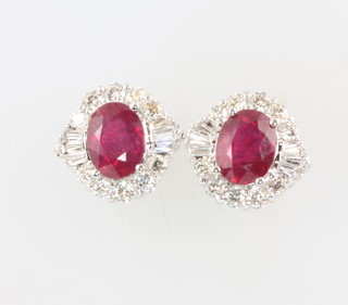 A pair of 18ct white gold oval treated ruby and diamond cluster earrings, the rubies 3.54ct, the tapered baguette and brilliant cut diamonds 1.04ct