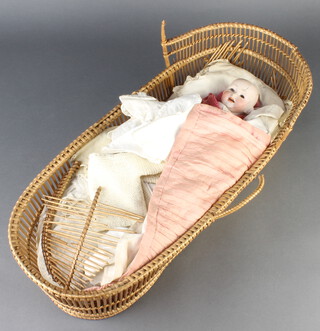 A German porcelain headed doll with open mouth, sleep eyes, head inscribed Made in Germany 152 (severely cracked and repaired) contained in a wicker crib (damaged) 17cm x 54cm x 23cm 