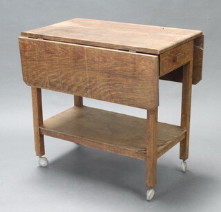 An oak drop flap tea trolley with drawer and undershelf on square legs and castors 70cm h x 71cm w x 46cm d (The trolley is faded and the undershelf veneer is lifting)