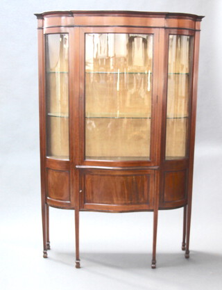 An Edwardian inlaid mahogany display cabinet of serpentine outline, the upper section enclosed by glazed panelled door, the base fitted a cupboard enclosed by a panelled door, raised on square tapered supports, spade feet 191cm h x 126cm w x 46cm d 