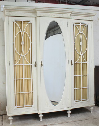 An Edwardian white painted breakfront triple wardrobe with moulded cornice, the centre fitted a cupboard with trays and drawers enclosed by an oval bevelled plate mirrored door flanked by cupboards enclosed by lattice work doors, raised on turned supports 214cm h x 191cm w x 65cm d 