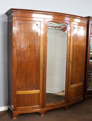 An Edwardian mahogany bow front triple wardrobe with moulded cornice, the interior fitted 3 trays above 3 short drawers with brass swan neck drop handles, enclosed by a bevelled plate mirror panelled door, flanked by a pair of panelled doors, raised on bracket feet 210cm h x 187cm w x 51cm d 