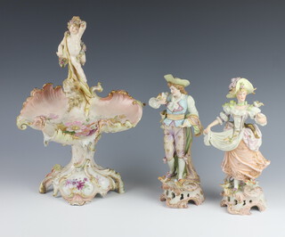 A pair of 19th Century German porcelain figures of standing lady and gentleman with birds 25cm h (both f and r), together with a scallop shaped centrepiece surmounted by a standing figure of a cherub (f and r) 
