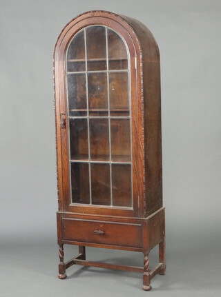 A 1930's oak arch shaped display cabinet, fitted shelves enclosed by a  lead glazed panelled door, the base fitted a drawer, raised on spiral turned supports with H framed stretcher 165cm h x 57cm w x 32cm d 