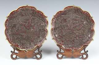 A pair of 19th Century brass mounted carved Cinnabar lacquer trays decorated with figures in a busy townscape, with carving and seal mark to base 20cm, on hardwood stands