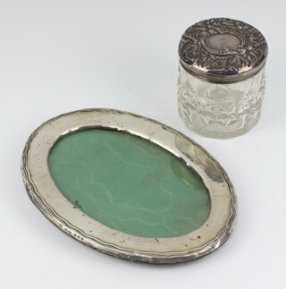 An oval silver photograph frame 17cm x 12cm, a cylindrical cut glass dressing table jar with silver lid (both have minor dents) 