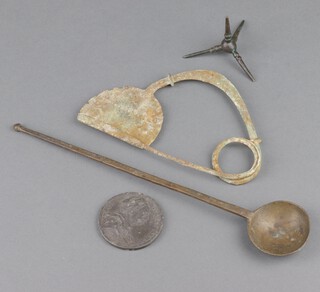 An Ancient Green or Roman bronze fibular brooch 16cm x 10cm, a bronze Caltrop 7cm, a bronze spoon 27cm and a token 