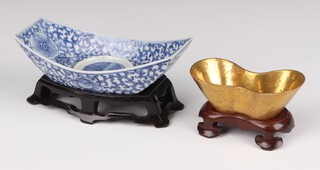 A gilt-decorated ingot-shaped vessel, Qing dynasty or later, the slanted sides rising to a straight rim, wood stand 3cm by 9cm together with a blue and white provincial ingot-shaped vessel, painted with two bats, musical chimes and peony scrolls, wood stand, 3.5 cm by 14.5cm long   (2)  