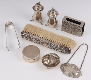 A pair of Georgian style silver pepperettes Birmingham 1904, a circular silver pill box with hinged lid (a/f), a pair of silver ice tongs, miniature silver salt and a silver whisky decanter label, 150 grams, together with a silver backed clothes brush and an Eastern white metal match box slip  