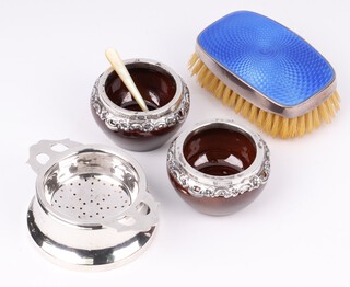 A silver and blue enamelled military style hair brush 8cm Birmingham 1927, a silver tea strainer and stand Birmingham 1936 (some dents) together with a pair of Edwardian brown glazed salts with embossed silver mounts Birmingham 1906 