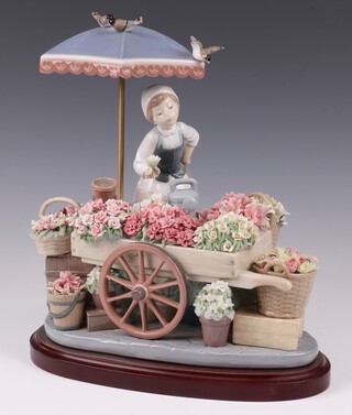 A Lladro figure group - The Flowers of The Season 1454, modelled as a flower girl with cart and baskets profusely decorated with encrusted flowers, beneath a parasol and with wooden base