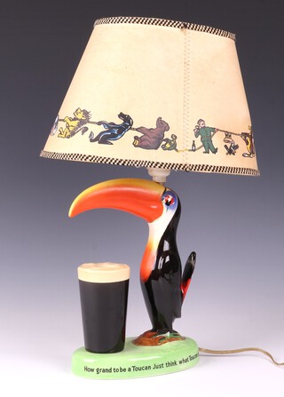 A Carlton Ware Guinness advertising table lamp in the form of a standing toucan with pint of Guinness, the base of the lamp marked "'How Grand To Be a Toucan Just think what Toucan do" and "If he can say as you can Guinness is good for you", with original shade featuring the Guinness characters in a tug-of-war, printed marks, 42cm high overall

