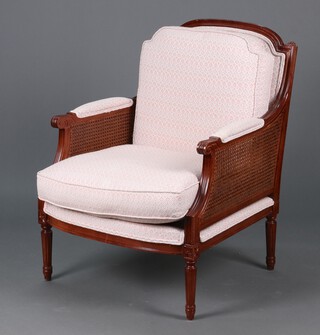 A carved mahogany Bergere style armchair upholstered in pink and white material with caned panels raised on turned and fluted supports